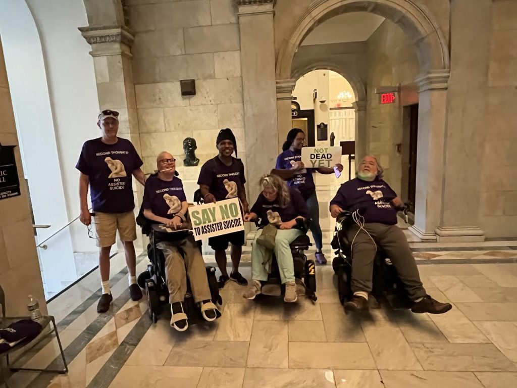 John Kelly and other advocates at the state house.  Signs say "Not Dead Yet" and Say No to Assisted Suicide.,  They are all wearing purple shirts that say Second Thoughts - No Assisted Suicide